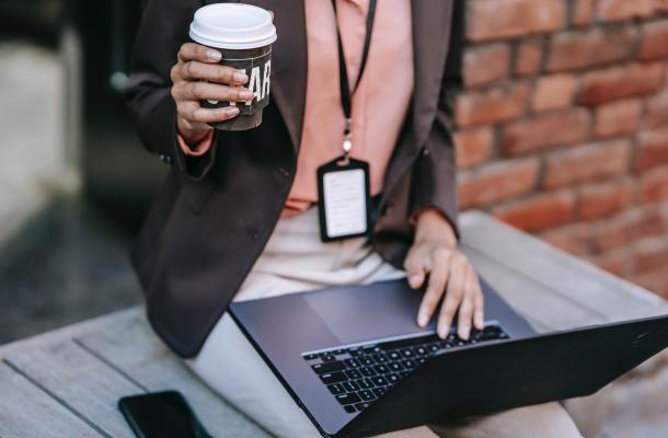 woman wearing a security badge holds a coffee while working on a laptop